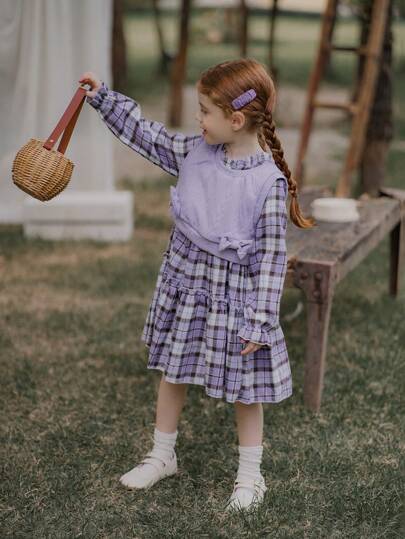 女孩的紫色蝴蝶结针织背心和紫色格子连衣裙，胸前有蝴蝶结装饰，看起来很可爱，紫色和格子的组合营造出温暖舒适的感觉，这款连衣裙有可爱的蝴蝶结细节，保持温暖舒适，适合秋冬季休闲、户外、日常穿着