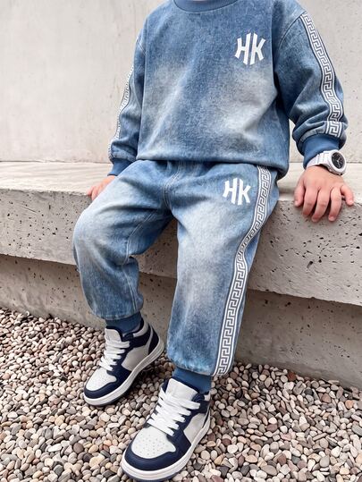 2-delat sweatshirtset för pojke med denimliknande textur, rundhalsad sweatshirt + långbyxor med mudd, ribbad halsringning, muddar och fåll, vitt HK-logotryck på vänster bröst, vitt mönstrat randtejp på yttersidan av ärmarna, enfärgade ribbade muddar på byxorna, samma mönstrade randtejp på byxornas sidor, vitt HK-tryck på låren
