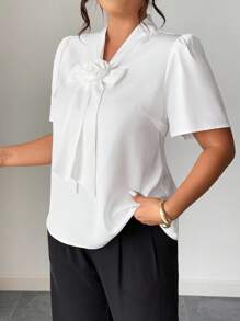 Elenzga 这款优雅的大码白色女式衬衫，领口饰有精致的蝴蝶结，并点缀着柔美的布艺花朵。这款别致的梭织上衣采用时尚的半泡泡袖设计，非常适合办公室或特殊场合穿着。