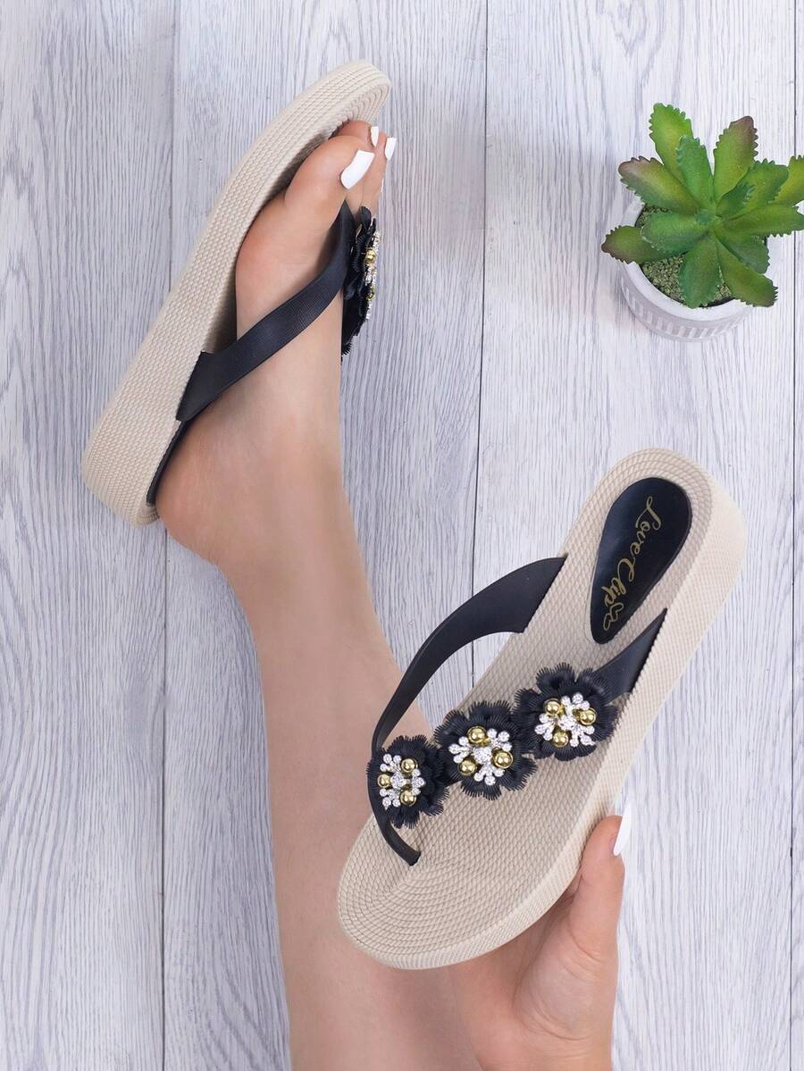 Sandalias tipo flip-flop de moda para mujer. La suela ofrece confort y suavidad a cada paso, mientras que las tiras negras están decoradas con tres elegantes flores adornadas con pedrería brillante en tonos dorado y blanco, dando un toque único y sofisticado. Su diseño ligero y cómodo es perfecto para llevar en el día a día, en la playa, vacaciones o simplemente para relajarte con estilo en casa. Las sandalias son fáciles de combinar con vestidos, shorts o jeans. - Negro - Ver 1