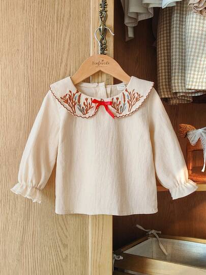 SHEIN Vintaside Kids Baby Girl Spring And Autumn Apricot-Colored Cotton And Linen Doll Neck Long-Sleeved Top Features A Ladylike Forest Style With A Sweet And Retro Charm. The Delicate Plant Embroidery Patterns On Both Sides Of The Doll Neck Are Decorated With Red Bows, And The Detailed Dog-Tooth Edges Make It Suitable For To School