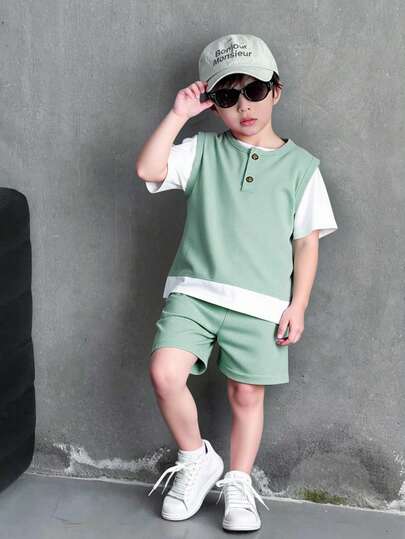 SHEIN Genkimix Kids 两件套儿童休闲套装 + 帽子，整体采用绿色针织面料。套装包含：上衣 - 仿二合一设计，绿色背心内搭白色短袖衬衫，格纹下摆打造层次感。下装 - 同色系绿色短裤，简约利落。配饰 - 同色系绿色渔夫帽，营造清新休闲的日常穿搭风格。