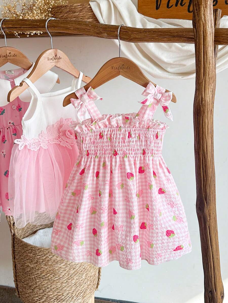 SHEIN Vintaside Kids 这款女童夏季粉色格子吊带连衣裙，以柔和的粉色格子为底色，点缀着新鲜草莓图案，可爱俏皮，散发着甜美乡村公主风情。连衣裙采用梭织面料，A字版型，肩带上的蝴蝶结装饰灵动可爱。适合草莓园拍照、校园活动、居家休闲、度假旅行以及参加派对等各种场合。