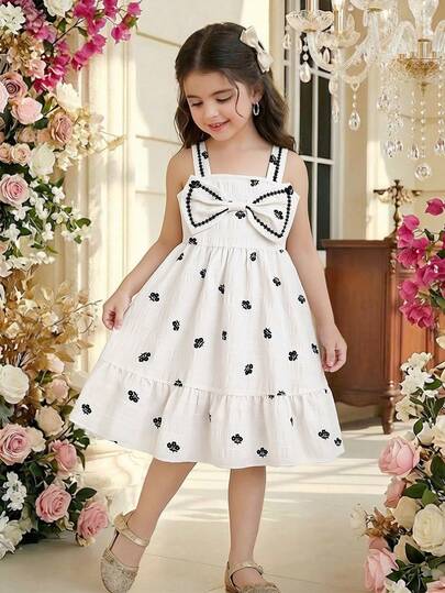 Young Girls' Sweet Forest Style Spaghetti Strap Dress, Made Of Light-Colored Fabric With Black Floral Embroidery Decor, Adorned With Large Bow Element On Chest, Ruffled Shoulder Straps And Layered Lace Hem, Loose And Flowing Silhouette, Suitable For Casual Outings, Family Gatherings, Lightweight And Comfortable