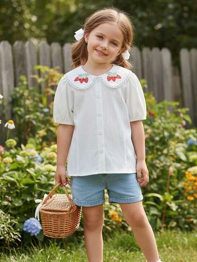 这款女童夏季短袖衬衫采用白色纹理面料，领口为彼得潘领设计，饰有精致的白色缝线，领子边缘点缀着白色刺绣草莓图案，领子两侧绣有红色草莓和绿色叶子，增添了一丝俏皮感。短袖采用泡泡袖设计，衬衫前襟有一排白色纽扣，整体风格甜美可爱，非常适合小女孩日常穿着。
