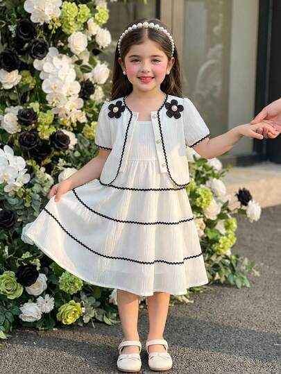 SHEIN Young Girls' Vintage French Style Cream White Spaghetti Strap Dress With Short Sleeve Cardigan Set. The Dress Is Made Of Vertical Striped Fabric With A Soft Touch, Featuring Layered Ruffled Hem With Black Wavy Trim. The Cardigan Has A Round Neckline Decorated With 3D Black Floral Trim And Black Wavy Trim. The Overall Design Is Fresh, Playful With A Touch Of Retro Sweetness. The Lightweight Dress And Thin Cardigan Are Suitable For Casual Outings, Park Walks, Or Family Gatherings In The Summer.