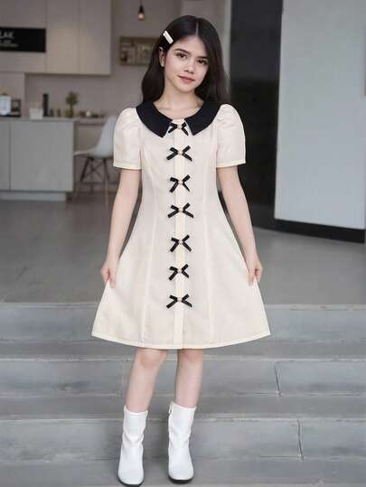 Elegante vestido de manga corta para niñas preadolescentes de talla media-grande en color albaricoque, con un diseño de cuello simple y manga corta, perfilado con líneas negras en el cuello y los puños para un aspecto limpio y refinado, decorado con un lazo negro en la parte delantera, manteniendo la elegancia vintage del vestido con lazo delantero mientras agrega un toque alegre con el lazo, en general un estilo elegante y minimalista, adecuado para ocasiones formales ligeras, reuniones, actuaciones y talla grande