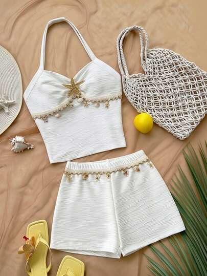 SHEIN 这款女童背心短裤套装，采用挂脖式设计，上衣饰有褶皱细节、星星和贝壳装饰，短裤正面则饰有贝壳和流苏。休闲简约，是春夏日常穿着的理想之选。