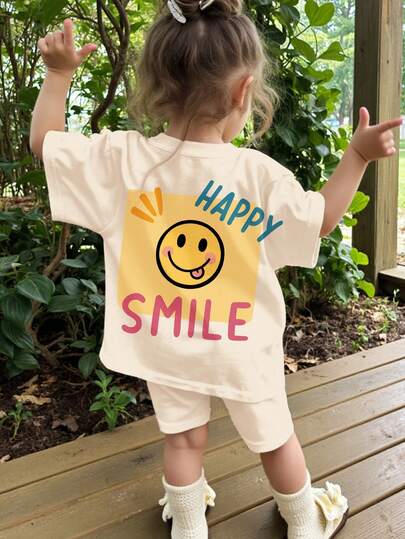 Bộ đồ gồm áo thun ngắn tay cổ tròn màu trắng kem, in hình mặt dễ thương, và quần short ôm sát dành cho bé gái, thích hợp mặc hàng ngày, dạo phố, đi nghỉ mát, dự tiệc, đi học, và các trang phục mùa xuân & hè.