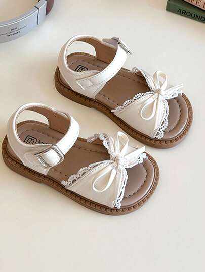 A Pair Of Popular White Children's Sandals With A White Strap And Metal Clasp On The Upper, A White Bow Trim And Lace Lace On The Front. The Style Is Sweet And Fresh, Belongs To The Summer Fashion Style, Suitable For Children's Daily, Holiday And Other Scenes, Suitable For 3-12 Years Old Children