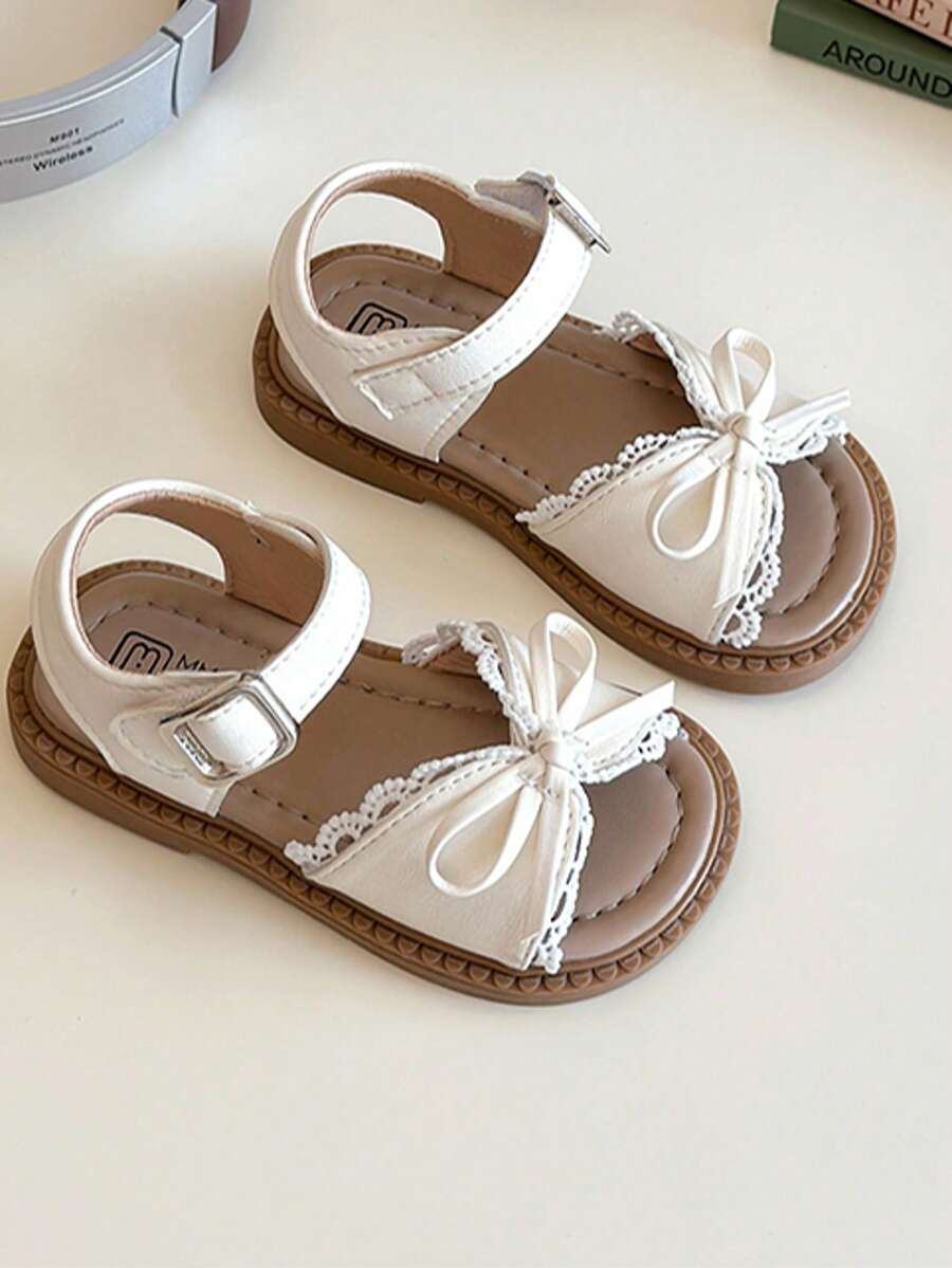 A Pair Of Popular White Children's Sandals With A White Strap And Metal Clasp On The Upper, A White Bow Trim And Lace Lace On The Front. The Style Is Sweet And Fresh, Belongs To The Summer Fashion Style, Suitable For Children's Daily, Holiday And Other Scenes, Suitable For 3-12 Years Old Children
