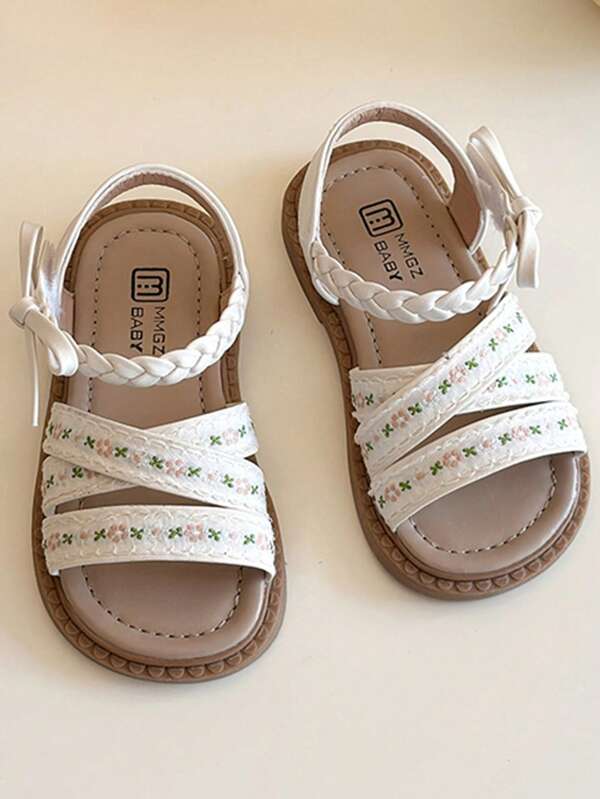 زوج من صنادل الأطفال الأبيض الأكثر مبيعًا في الصيف ذو تصميم شريطي متضفر وحزام متقاطع مزين بطرز الزهور الصغيرة، مع فيونكة مزخرفة عند الكاحل وبنعل بني. النمط عذب وطازج، ينتمي إلى أزياء الصيف، مناسب للأطفال من عمر 3-12 سنة لاستخدامهم اليومي والعطلات وغيرها من المناسبات