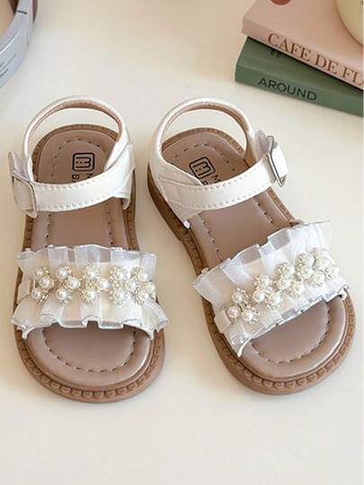 A Pair Of Best-Selling White Children's Sandals With A White Strap And Metal Buckle Design, A White Pleated Front Embellished With Pearls. The Style Is Sweet And Elegant, Belongs To The Summer Fashion Style, Suitable For Children's Daily, Holiday And Other Scenes, Suitable For 3-12 Years Old Children