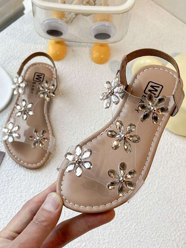 Ein Paar der Kinder-/Mädchen-Sandalen. Das Obermaterial des Schuhs besteht aus transparentem Material und ist mit mehreren Strass-Blumen verziert. Die Blumenformen sind elegant und glänzend. Ein hellbrauner Schnallenverschluss befindet sich am Knöchel. Der Gesamtstil ist süß und verspielt, voller Prinzessinnen-Charme. Es ist geeignet für Mädchen im Alter von 3 bis 12 Jahren, um sie bei Sommerfeiern, Fotoshootings und anderen Anlässen zu tragen und ihre Süße und Verspirtheit zu zeigen.