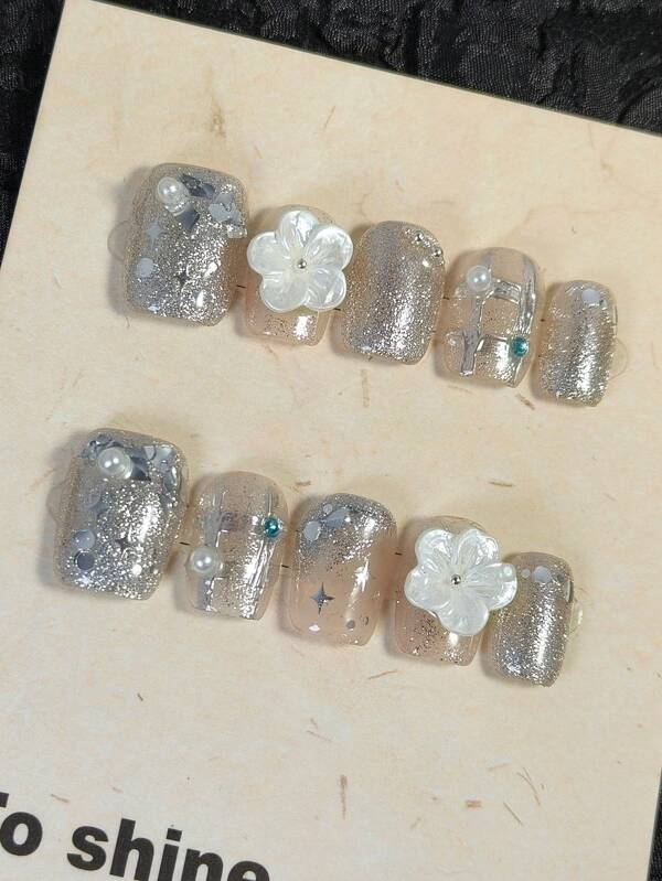 Un set de 10 ongles courts et carrés, élégants, présentant un dégradé argenté peint à la main dans le style Y2K, des paillettes fines scintillantes, de grandes sequins brillants, des formes de diamants roses miroirs, des étoiles à quatre pointes, des fleurs blanches tridimensionnelles, des perles, des boules d'acier et des diamants bleus.