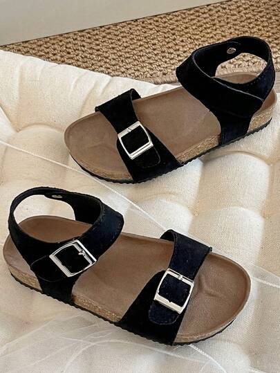 Ett par barnsandaler/flicksandaler från 2025 med svart matt ovandel, dubbel rem och metallspänne samt ett omlottband vid vristen, vilket ger en enkel och trendig stil.