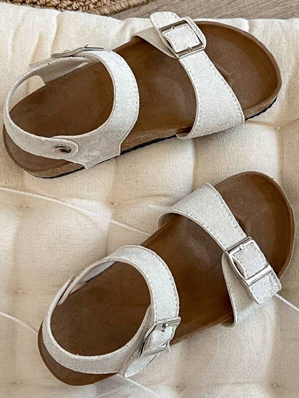 Un par de sandalias para niñas 2025, con Top mate beige, con diseño de doble tira y hebilla de metal, y tira envolvente en el tobillo, presentando un estilo sencillo y de moda.