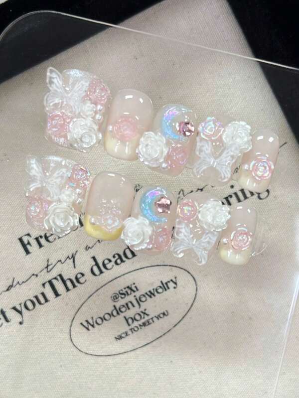 Cet ensemble de nail art comprend 10 ongles courts carrés faits main, ornés de roses roses aurorales, de strass, de camélias blancs, de perles, de strass en forme de papillon et de designs de strass en forme de lune et d'étoile, le tout dégageant un brillant .