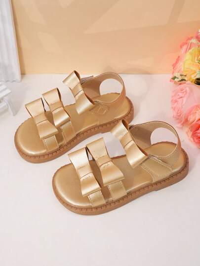一双女童金色蝴蝶结多带凉鞋，鞋面采用亮面人造革，质感精致。魔术贴和多带设计可调节松紧度，露趾低帮款式，平底设计，甜美又时尚，适合夏季生日派对和节日庆典。