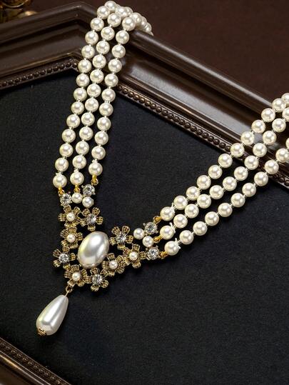A Multi-Layered Necklace With Rounded, Faux Pearl Chains That Create A Graceful, Vintage-Inspired Look. The Necklace Features Engraved Details Surrounding The Plump, Faux Pearls, And A Dangling Teardrop-Shaped Pearl Pendant That Adds A Delicate, Soft Touch. The Vintage-Luxe And Gentle Elegance Are Seamlessly Blended.
