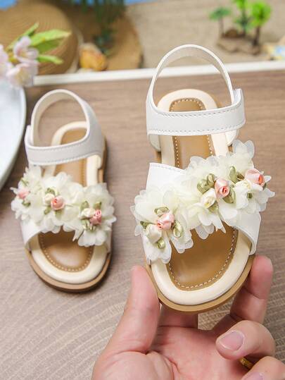 Giày sandal đế bằng hoa trắng mới cho bé gái mùa xuân/hè 2026, kiểu công chúa với hoa 3D bằng sợi len, đế mềm mại, thoải mái, giày La Mã đa năng cho trẻ em.