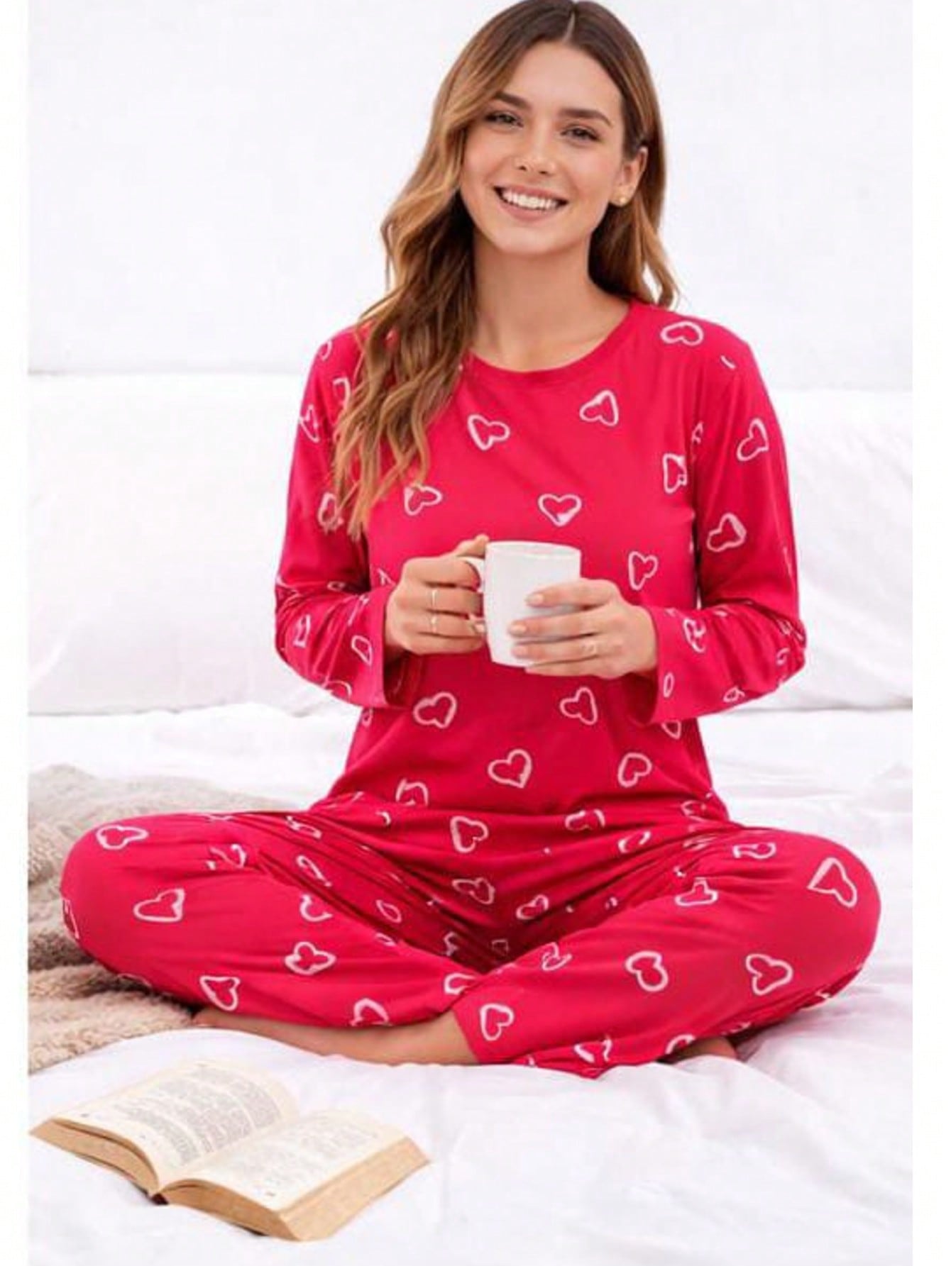 Modelo sentada na cama segurando uma xícara, vestindo pijama longo canelado de corações