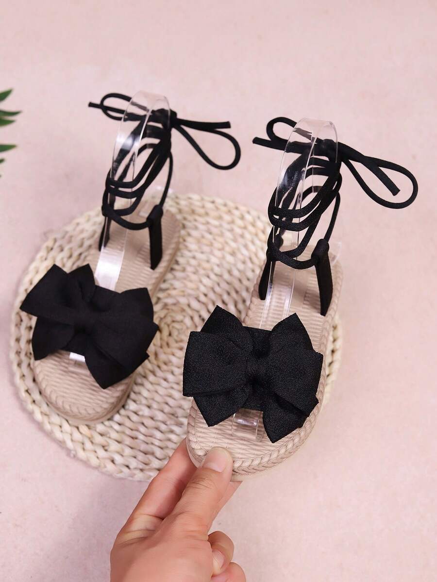 1 paire de sandales de plage plates, couleur unie en tissu doux avec grand nœud décoratif, bride de cheville élastique, bout rond, semelle flexible antidérapante, style romain mignon et à la mode pour filles, convient pour le quotidien, la plage et le campus au printemps et en été - Noir - Voir 1