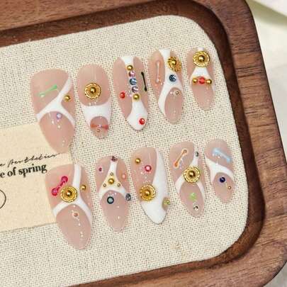 10 móng tay giả thủ công, với họa tiết đường kẻ hình học màu trắng bất đối xứng làm nền, được trang trí bằng các hạt cườm, đá quý và điểm nhấn kim loại vàng nhiều màu sắc, mang phong cách retro-futuristic và năng động, ngọt ngào và cá tính, rất riêng biệt.