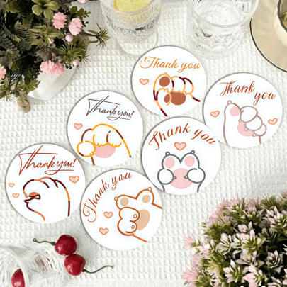 Dessous de verre thématique de Thanksgiving avec motifs de pattes et gestes de la main, avec un dessin au trait simplifié de jolies empreintes de pattes et de gestes de la main anthropomorphiques, associés à un texte "Merci" et des décorations de cœur, utilisant des couleurs douces à faible saturation et un style minimaliste de dessin au trait, transmettant une atmosphère douce et chaleureuse de Thanksgiving