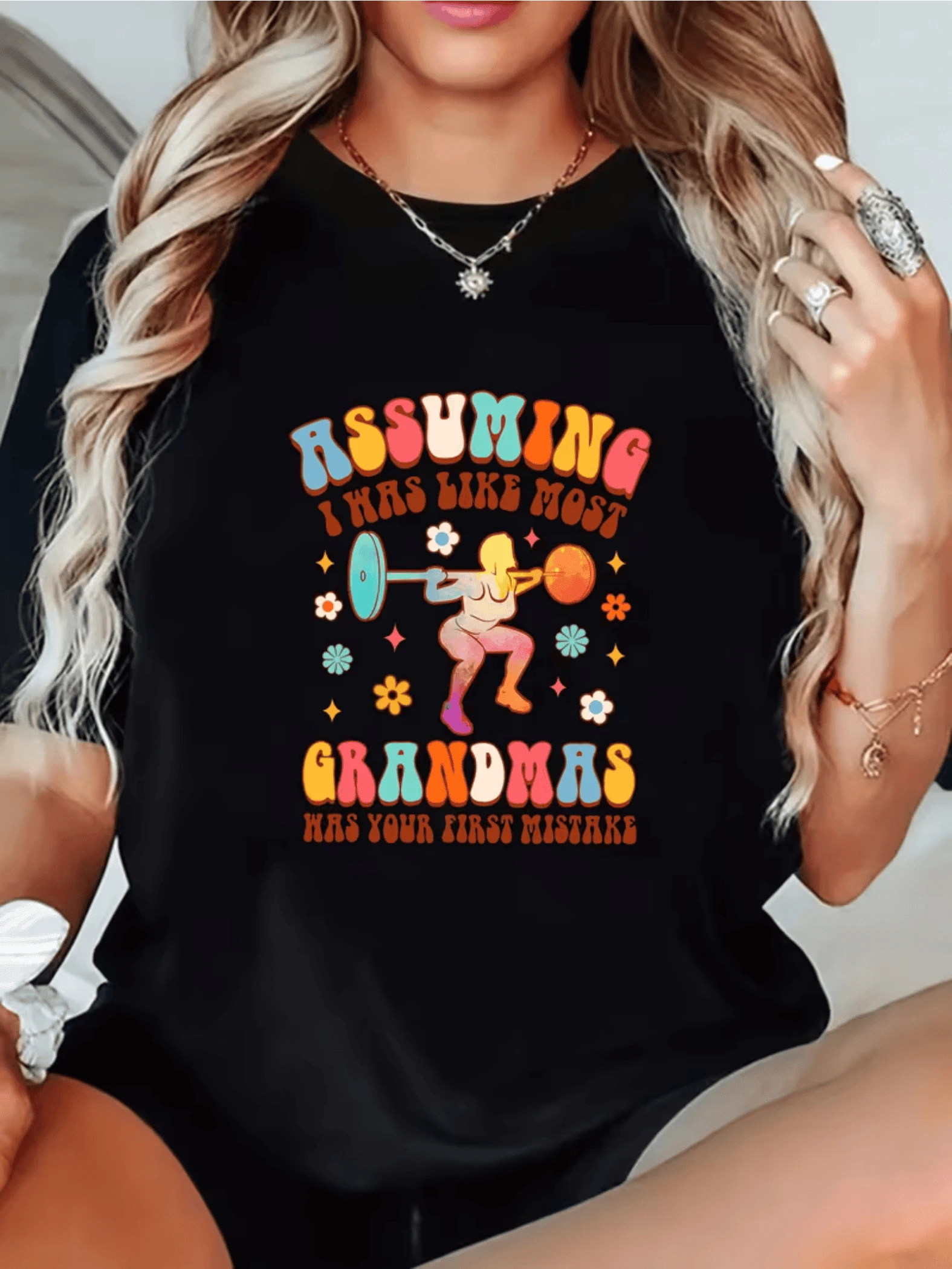 Colorful Illustration Depicts A Strong Grandmother Lifting Weights Surrounded By Small Flowers, Symbolizing Energy, Joy, And Feminine Confidence. Comfortable, Modern, Casual Women's T-Shirt For Everyday Wear. - 黑色 - 查看 1