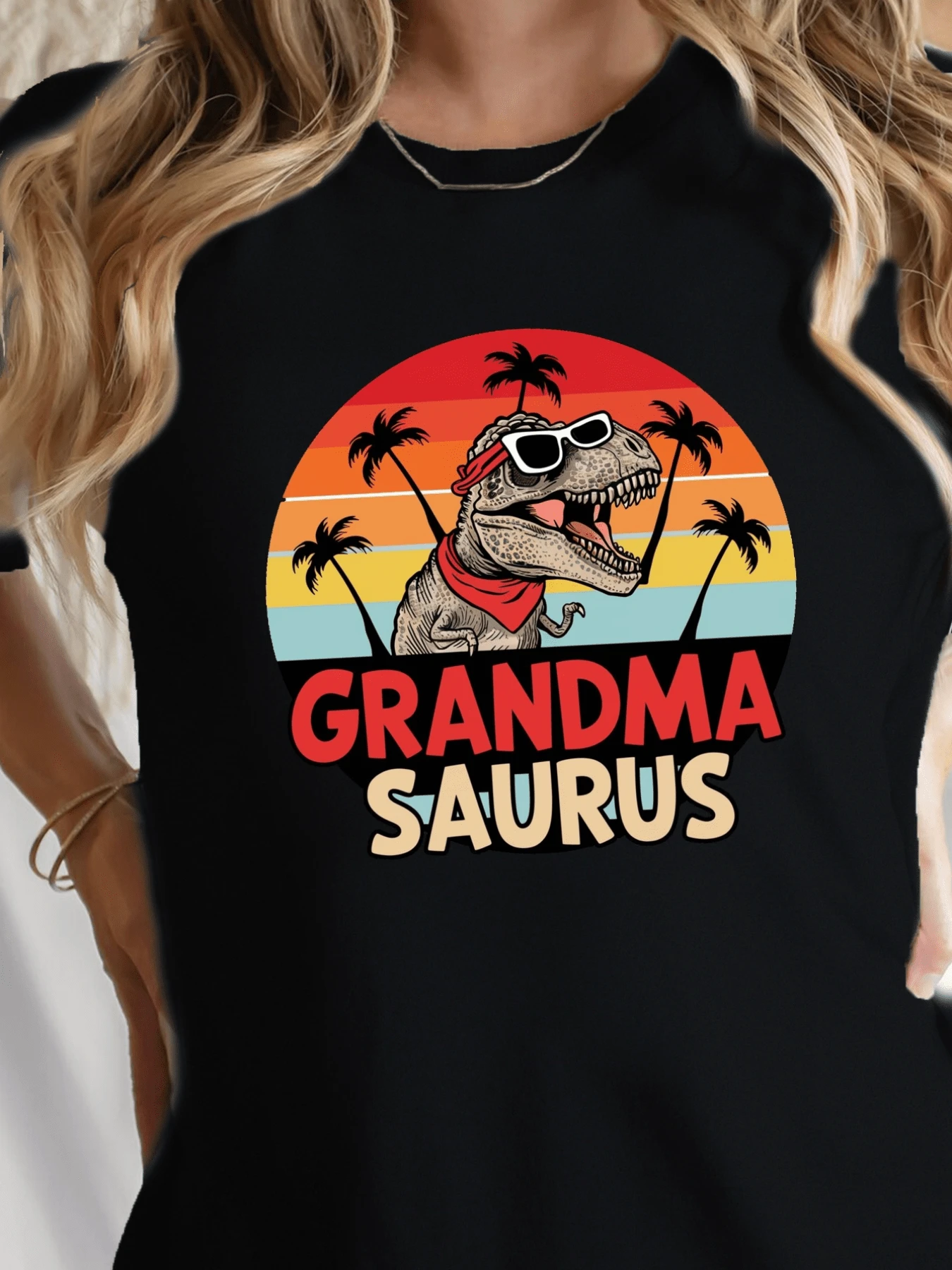 Smiling Tyrannosaurus Rex With Red Sunglasses And A Bandana Celebrating Among Palm Trees Against A Multicolored Sunset Symbolizes The Joyful Strength And Vitality Of An Adventurous Grandmother. A Modern And Fun Casual Women's T-Shirt. - 黑色 - 查看 1